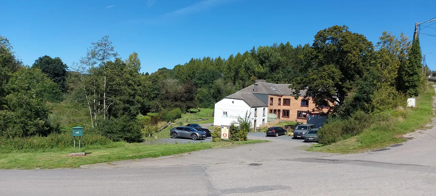 Vue sur la maison du Vieux Moulin. Il y a des arbres et des voitures devant ainsi qu'un ciel bleu.